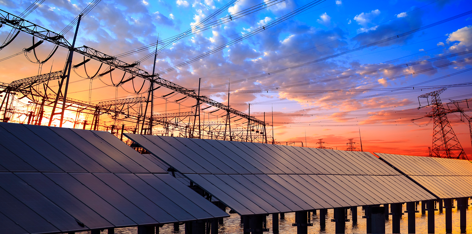 Solar panel array in front of high-voltage transmission towers at sunset