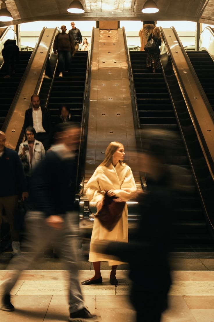 A woman stands still at the base of golden escalators while commuters blur past